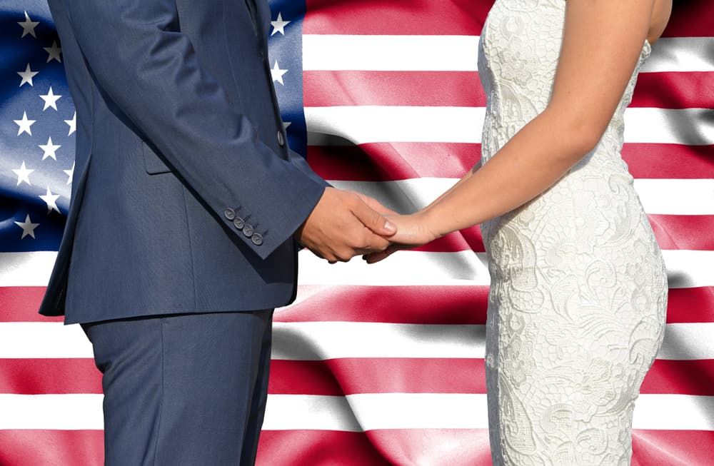 Casal em trajes de casamento de mãos dadas em frente a uma bandeira americana, simbolizando a comprovação de um casamento de boa-fé para o processo de imigração e green card dos EUA.