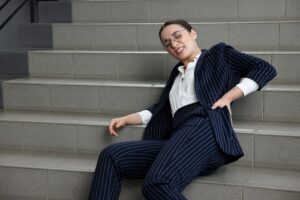 Businesswoman in a pinstripe suit sitting on stairs in visible pain after a fall, holding her lower back with a pained expression.