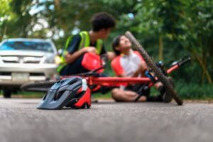 Bicycle accident scene with a helmet in the foreground and two injured cyclists sitting on the road, a car visible in the background.