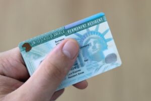 Hand holding a United States permanent resident card (green card), showing part of the front with the Statue of Liberty design visible.