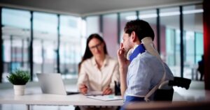 Injured man wearing a neck brace sitting in a wheelchair during a legal consultation with a female lawyer in a modern office.
