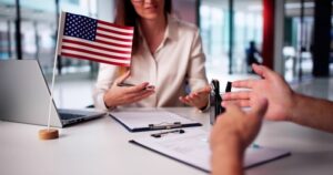 Close-up of a business meeting in a modern office. A small American flag is on the table beside documents, a pen, and a laptop, while a woman and another person gesture and discuss paperwork.