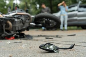 Close-up of broken motorcycle mirror on the road after an accident, with a motorcycle lying on its side in the background and two distressed individuals near a damaged car.
