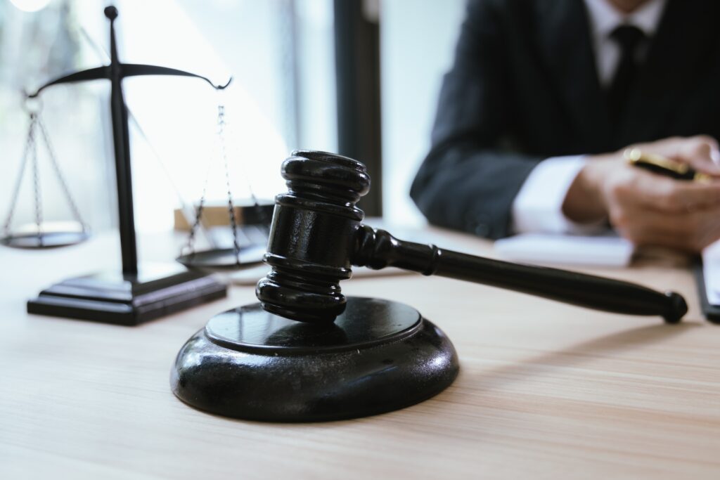 Judge&rsquo;s gavel on desk with scales of justice and attorney in background during legal proceedings.