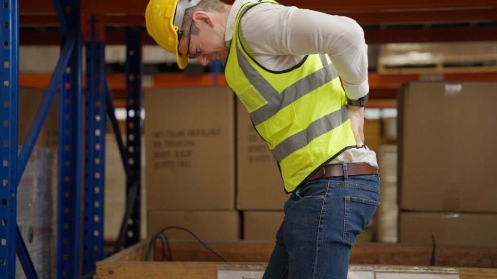 Warehouse worker wearing safety vest and hard hat experiencing lower back pain after workplace injury. Warehouse worker wearing safety vest and hard hat experiencing lower back pain after workplace injury.
