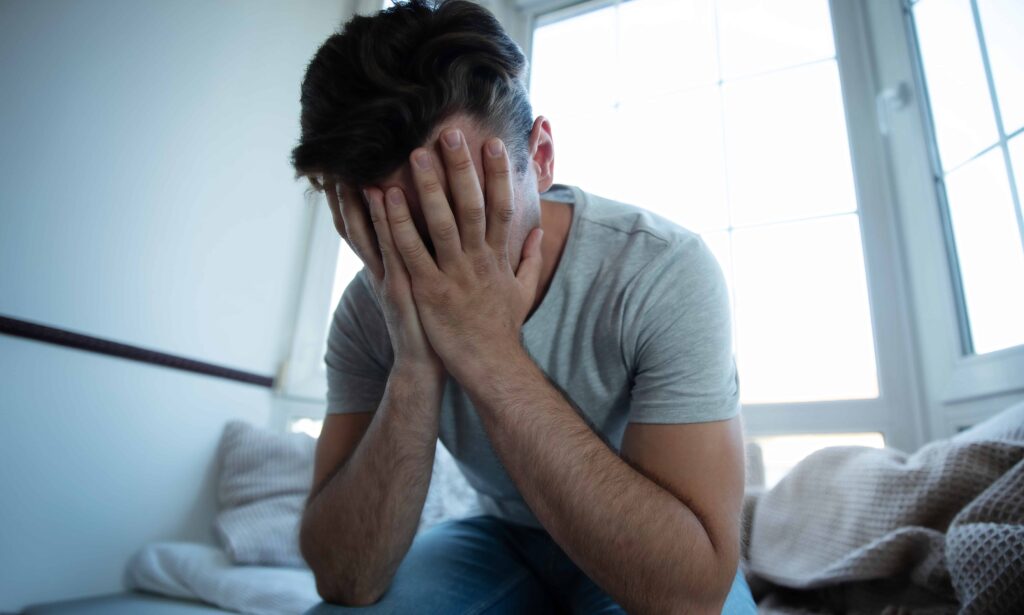 Stressed man sitting on bed with head in hands, showing emotional distress or mental health struggle.