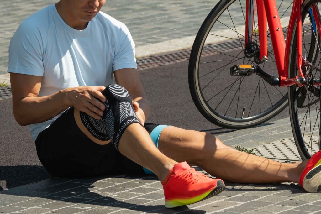 Cyclist sitting on pavement adjusting knee brace after bicycle accident or knee injury.