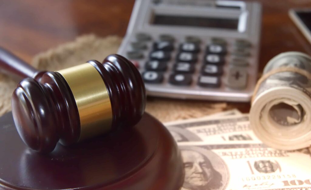 Judge’s gavel resting on U.S. dollar bills beside a calculator, symbolizing legal costs, settlements, or financial damages. Judge’s gavel resting on U.S. dollar bills beside a calculator, symbolizing legal costs, settlements, or financial damages.